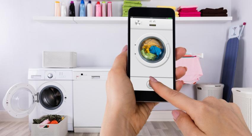 Photo showing hands of person holding smart phone. Smart phone shows photo of a washing machine. Background is laundry room.